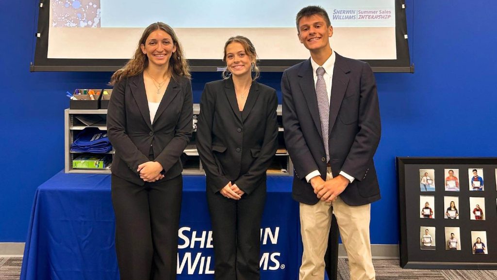 A photo of three people standing in front of a table and display screen.